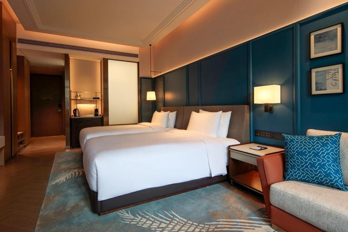 A hotel room with a double bed, a sofa, a nightstand, a lamp, and artwork on the wall.