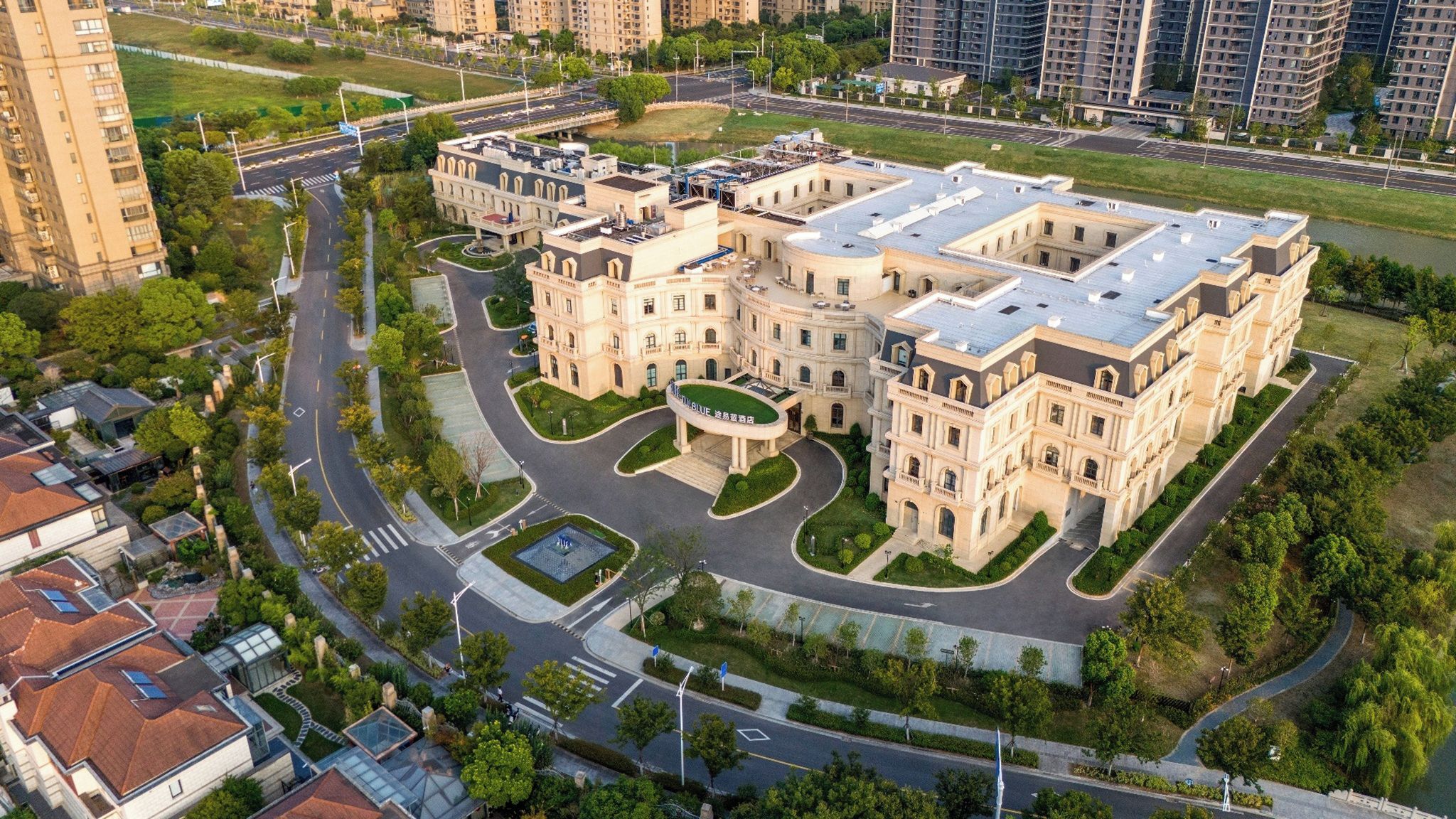 A multi-story hotel with a central courtyard, surrounded by greenery and adjacent to a road.