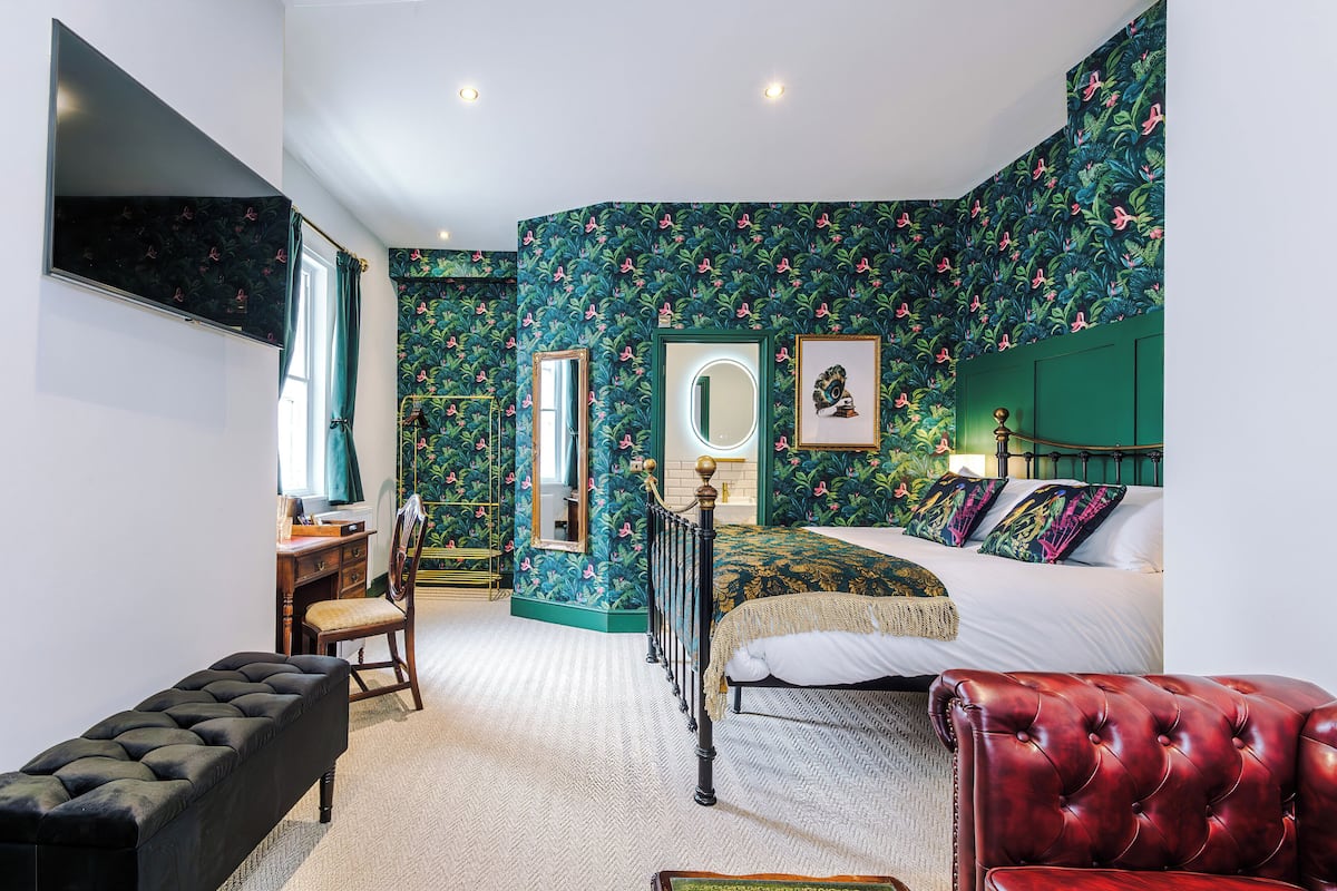 A bedroom with a bed, a bench, a desk, a chair, a mirror, and a wall with a floral pattern.