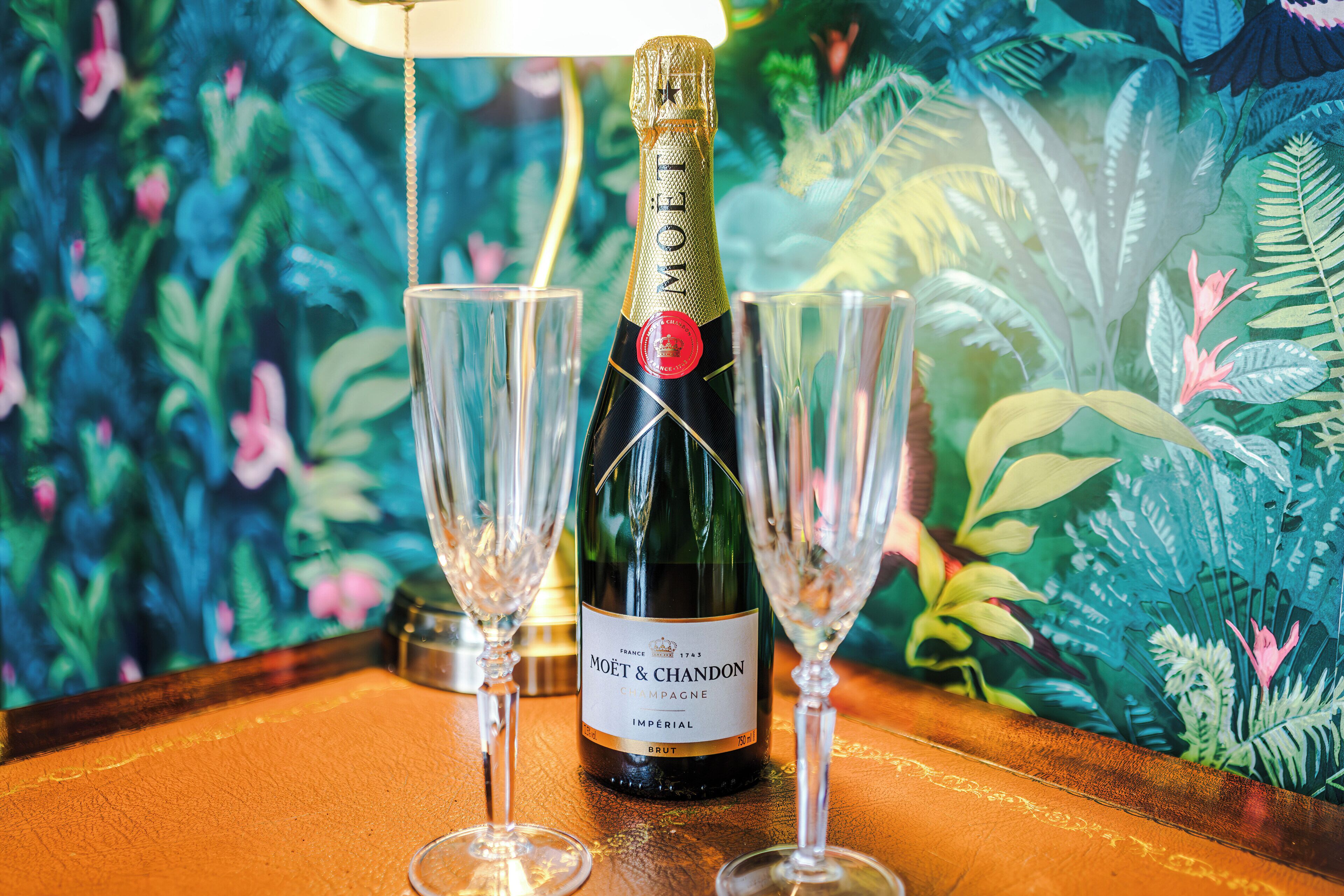 A bottle of Moet & Chandon champagne with two glasses on a wooden table against a tropical wallpaper background.