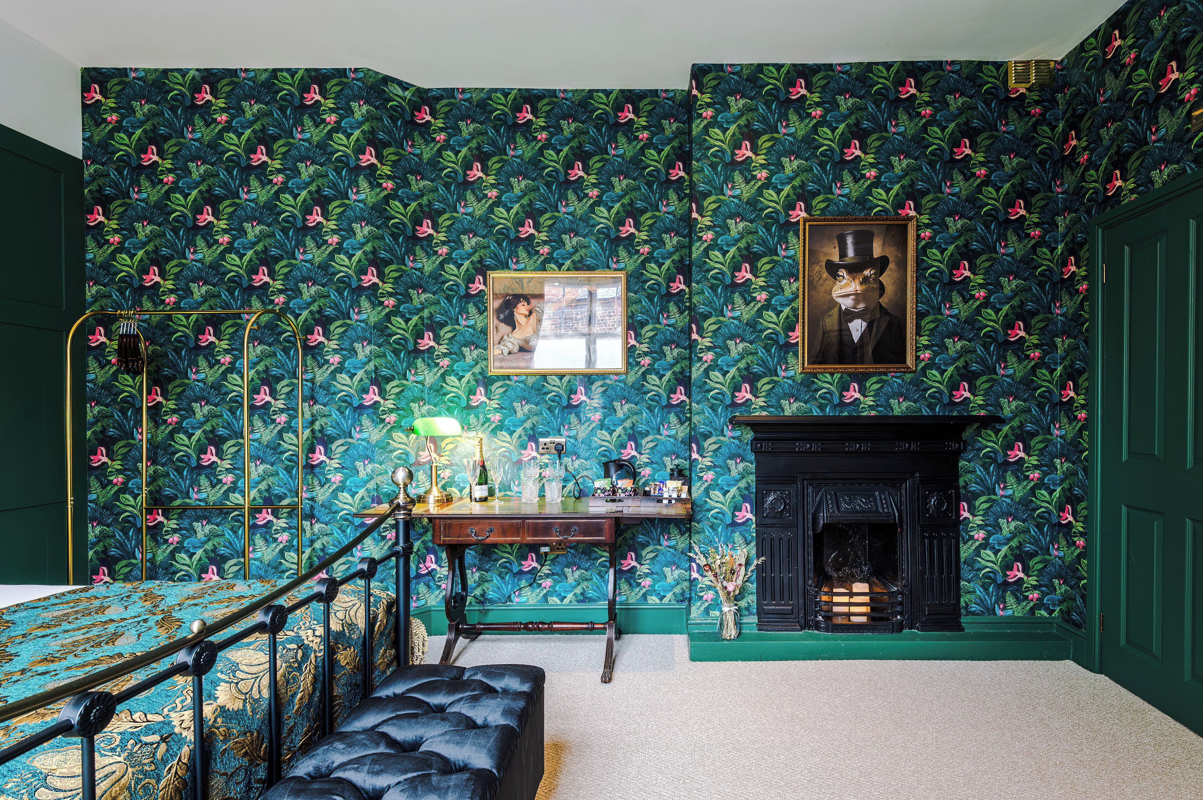 Une pièce avec un papier peint à motifs, une cheminée, un bureau avec une lampe et un tableau encadré au mur.
