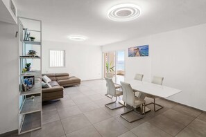 A modern living room with a sofa, a dining table with chairs, a bookshelf, and a large window. - Stunning Sea View 3BDR With Terrace By Isrentals (Netanya)