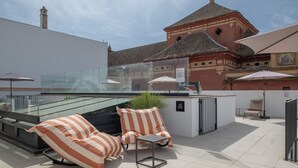 Een terras op het dak met twee gestreepte lounge fauteuils, een kleine tafel en een glazen hekwerk met uitzicht op een historisch gebouw met een pannendak.
