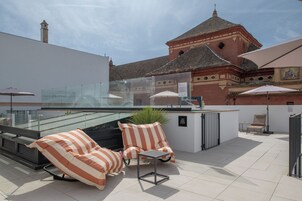 A rooftop terrace with two striped lounge chairs, a small table, and a glass balustrade overlooking a historic building with a tiled roof.