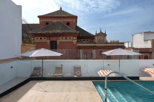 Una piscina en la azotea con sillas de descanso y sombrillas, un edificio con techo de tejas al fondo.
