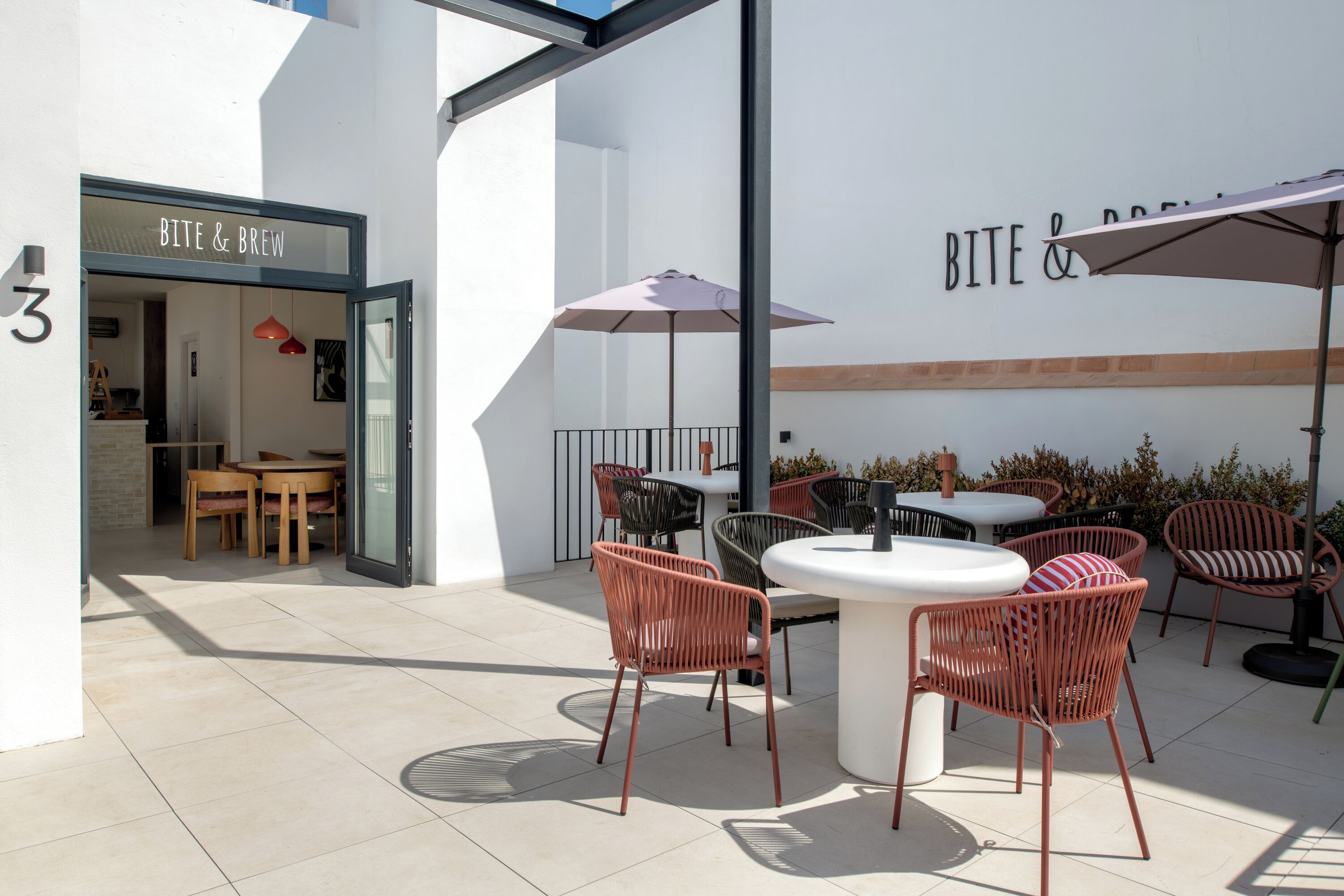 Ein Außenbereich mit Tischen, Stühlen und einem Sonnenschirm, sowie ein Gebäudeeingang mit der Beschriftung 'BITE & BREW'.