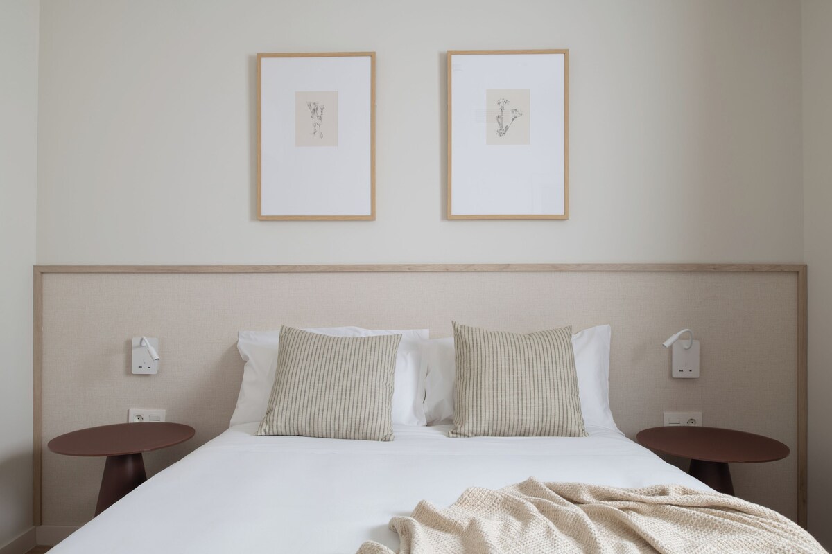 Una habitación de hotel con una cama, dos mesitas de noche, un cabecero y dos cuadros enmarcados en la pared.