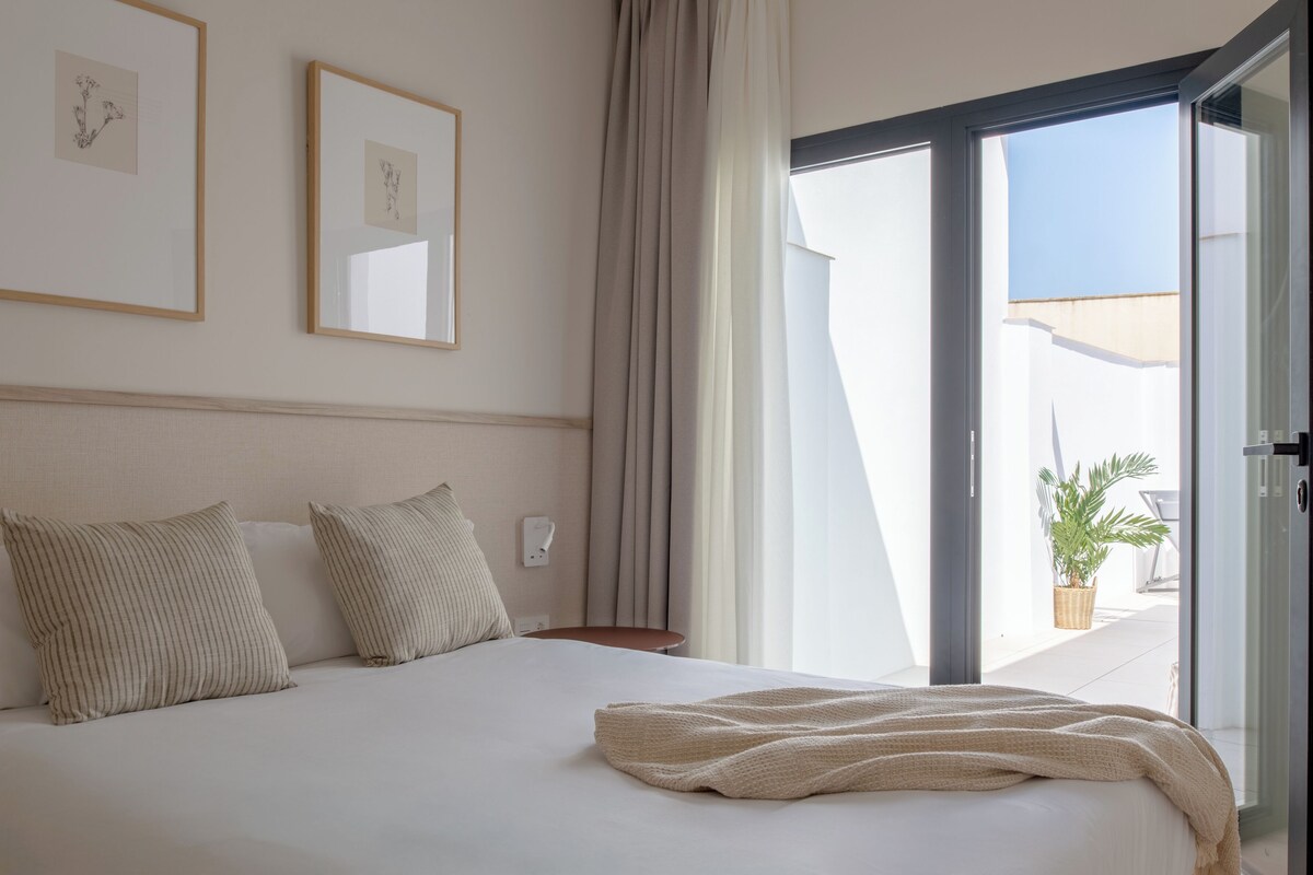 A bedroom with a bed, pillows, a bedside table, a window with curtains, and framed artwork on the wall.