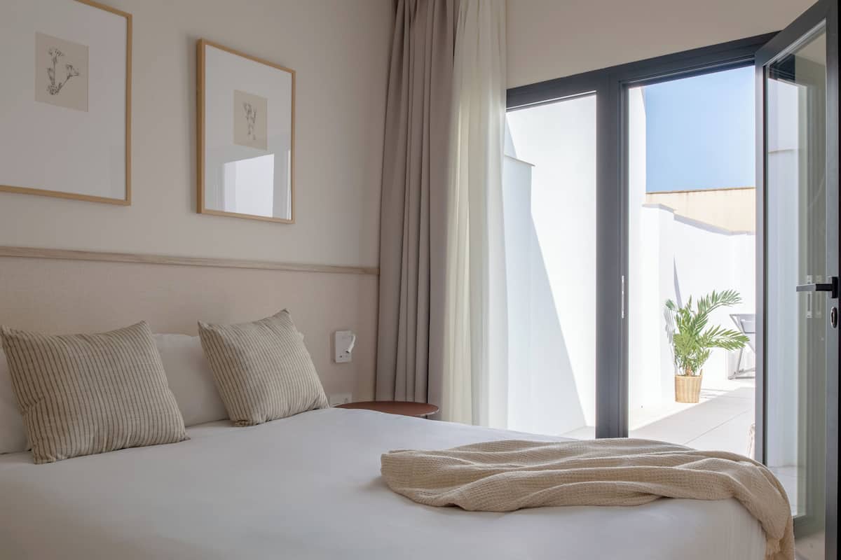 A bedroom with a bed, pillows, a bedside table, a window with curtains, and framed artwork on the wall.