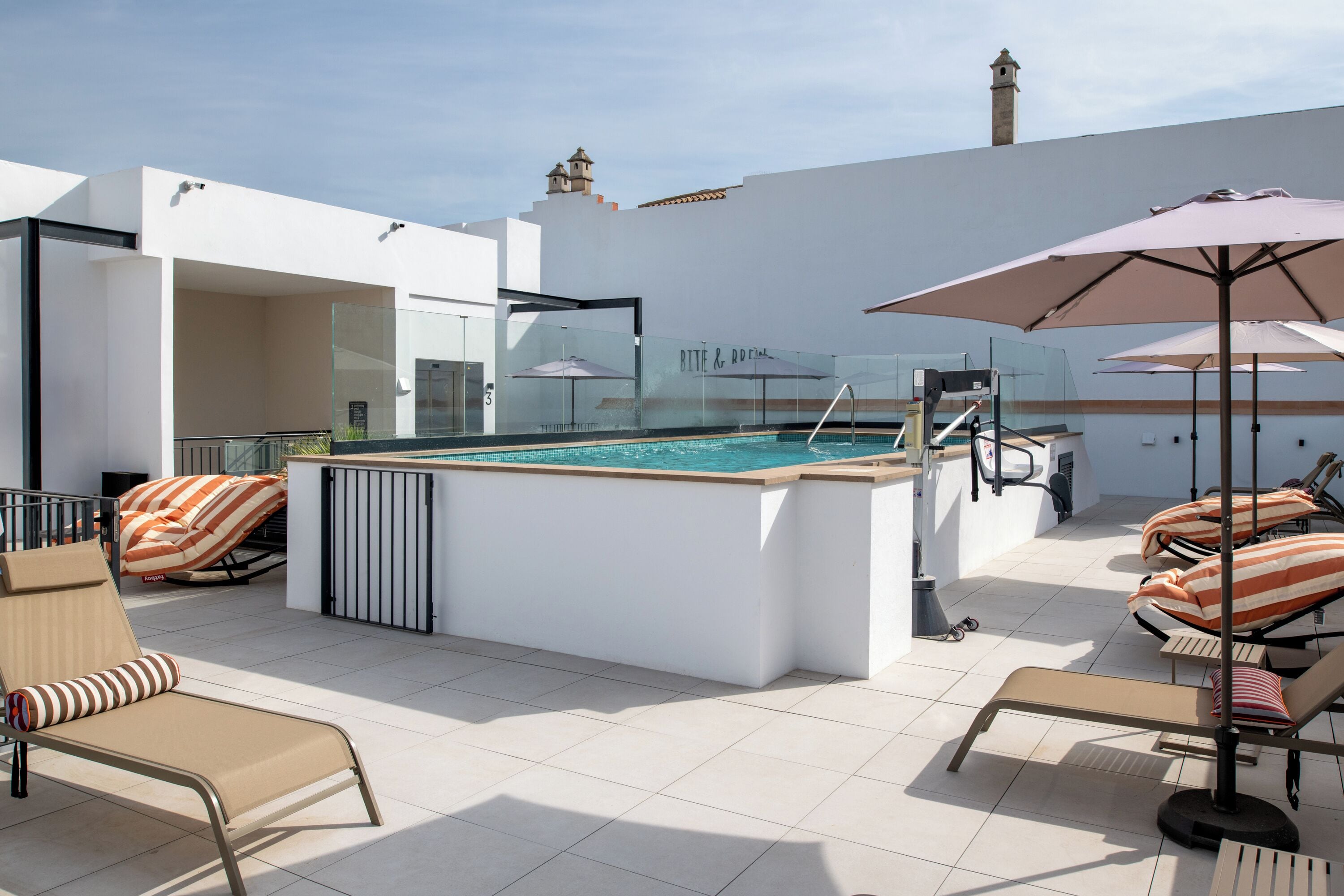 A rooftop pool area with lounge chairs, umbrellas, and a glass-enclosed pool.