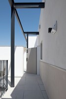 Un estrecho pasillo exterior con una reja de metal, una pared blanca y un cielo azul despejado.