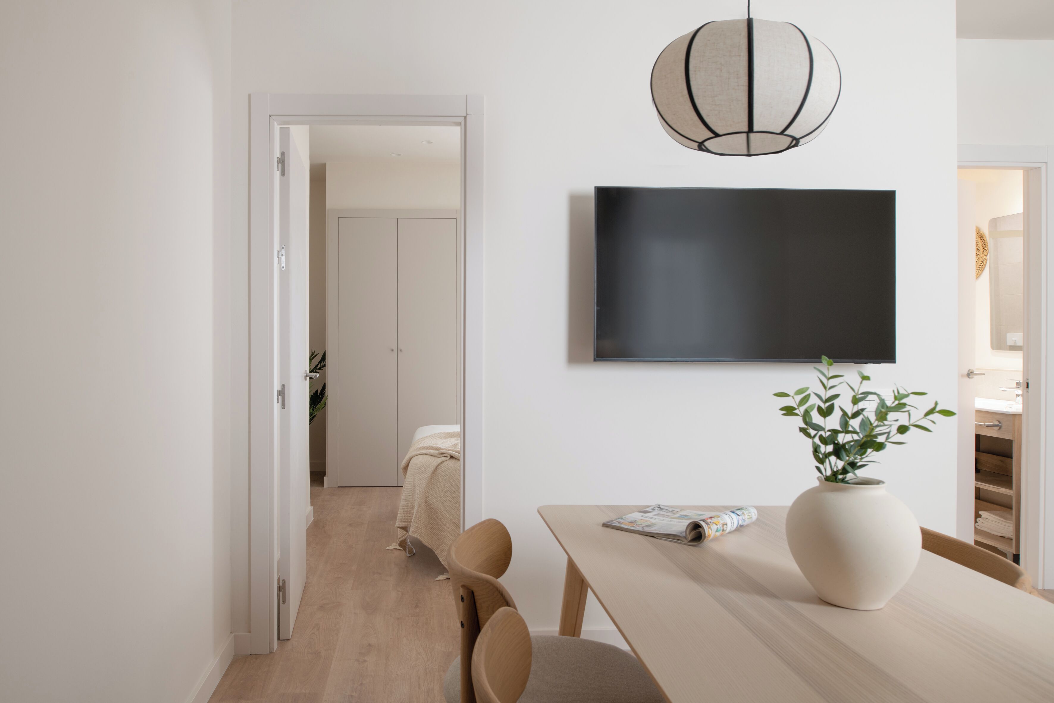 Un comedor moderno con una mesa de madera, sillas, un florero con planta y un televisor montado.