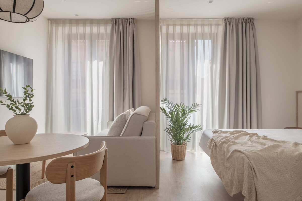 A modern hotel room with a round dining table, a sofa, a bed, and a potted plant.