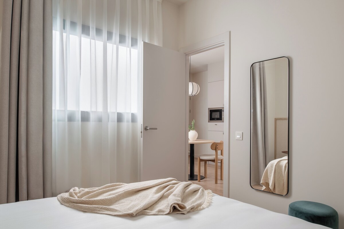 A bedroom with a bed, a bedside mirror, a window with curtains, and a view into another room.