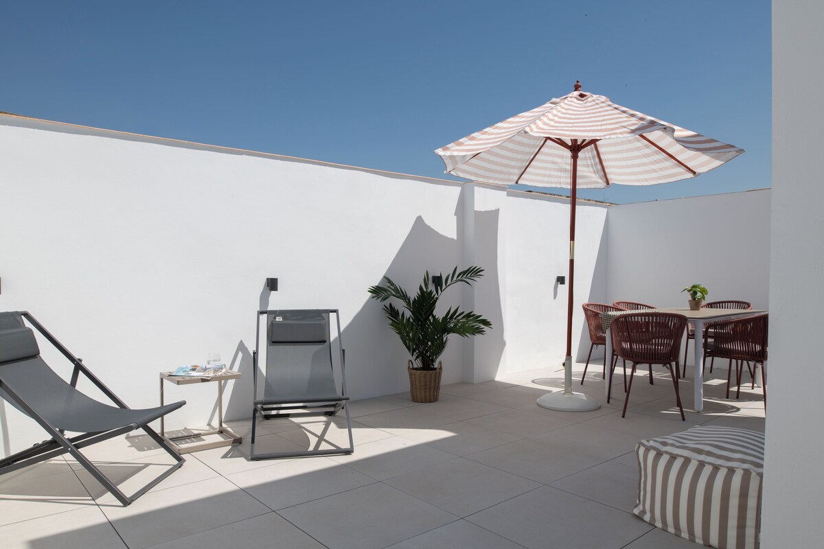 A sunlit outdoor patio with a striped umbrella, lounge chairs, a dining table with chairs, and a potted plant.
