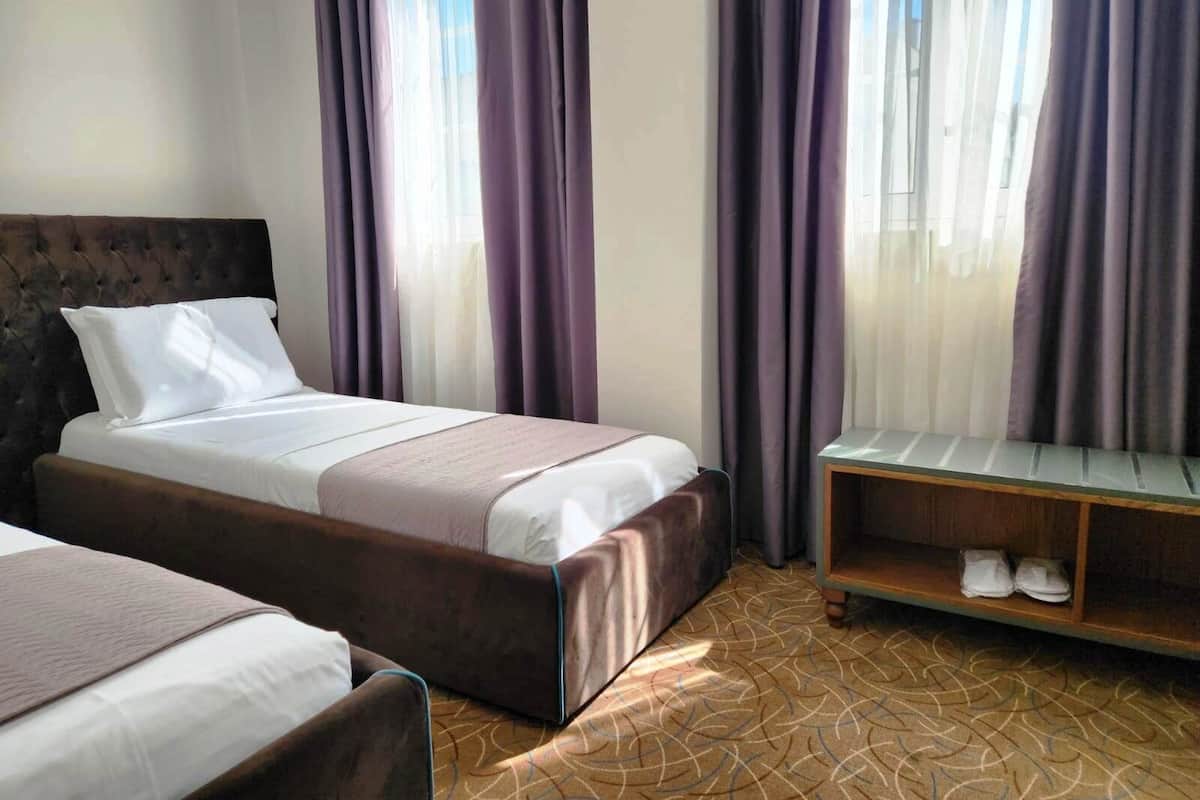 Habitación de hotel con dos camas, una mesita de noche de madera y una ventana con cortinas moradas.
