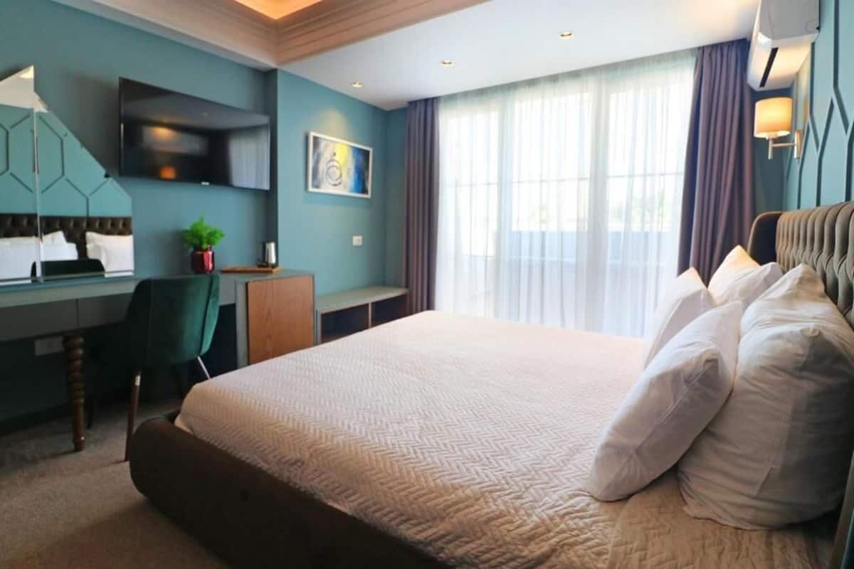 Habitación de hotel con una cama grande, un escritorio con silla, un televisor montado en la pared y una ventana con cortinas.