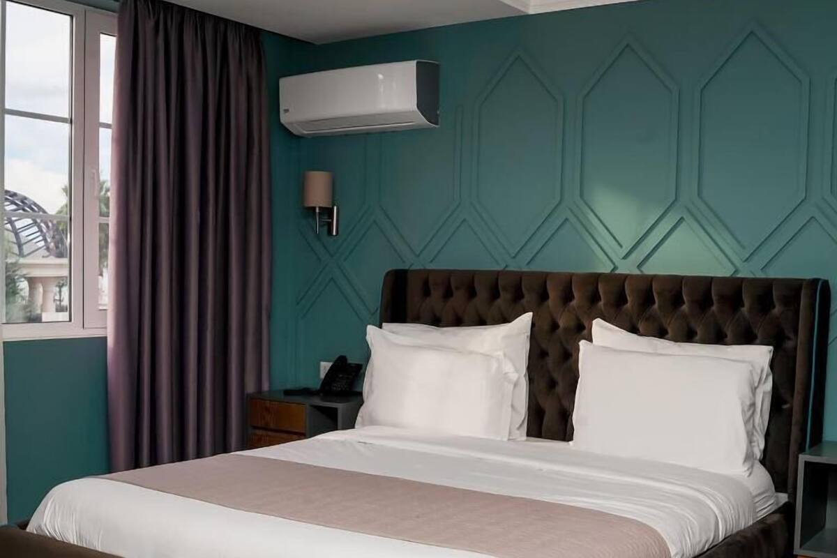 Habitación de hotel con una cama grande, una mesita de noche, una lámpara y una ventana con cortinas.