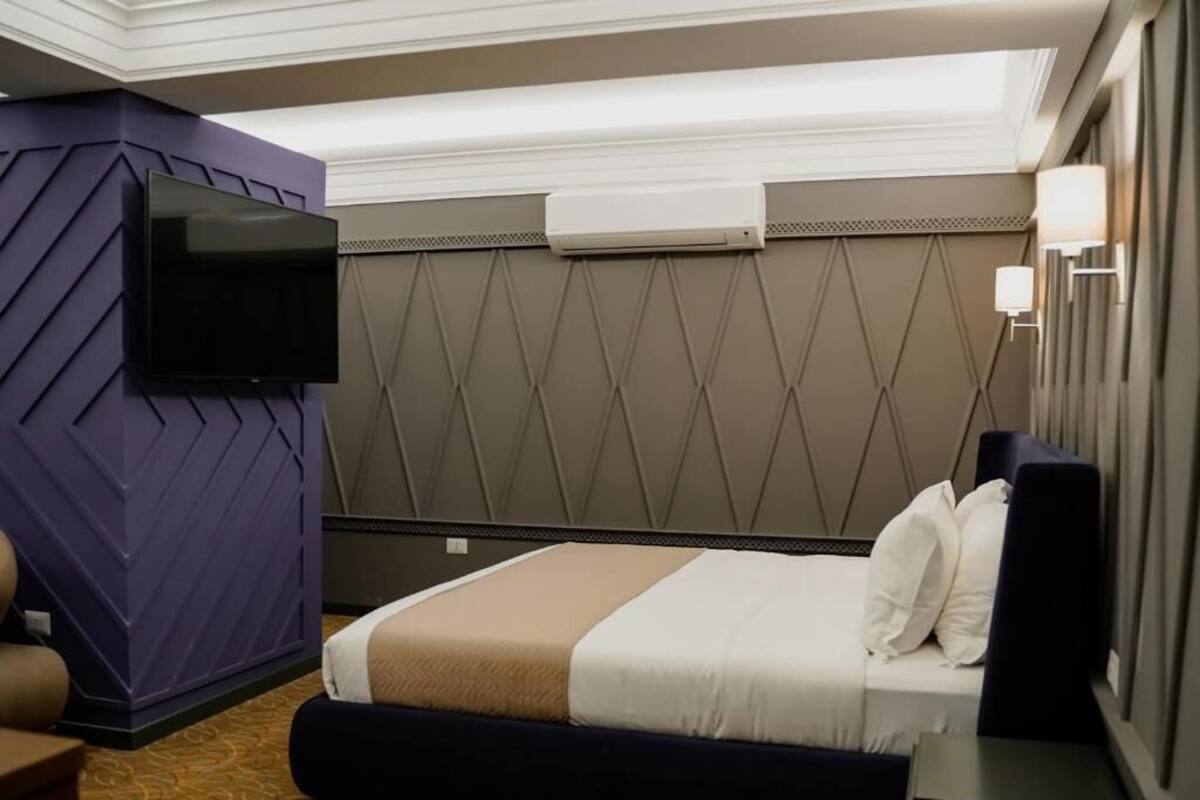 Habitación de hotel con cama, televisor de pantalla plana montado en la pared, mesita de noche y aire acondicionado mural.