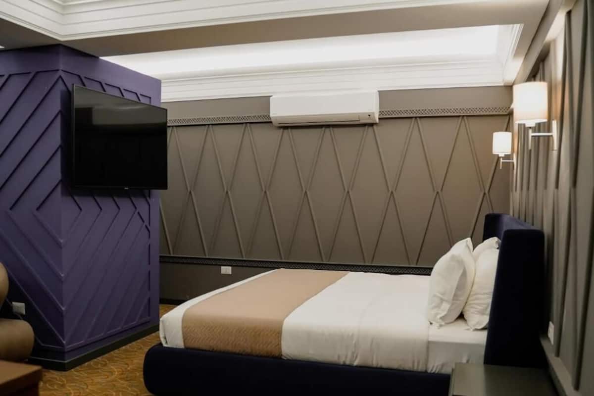 A hotel room with a bed, a flat-screen TV mounted on the wall, a bedside table, and a wall-mounted air conditioning unit.