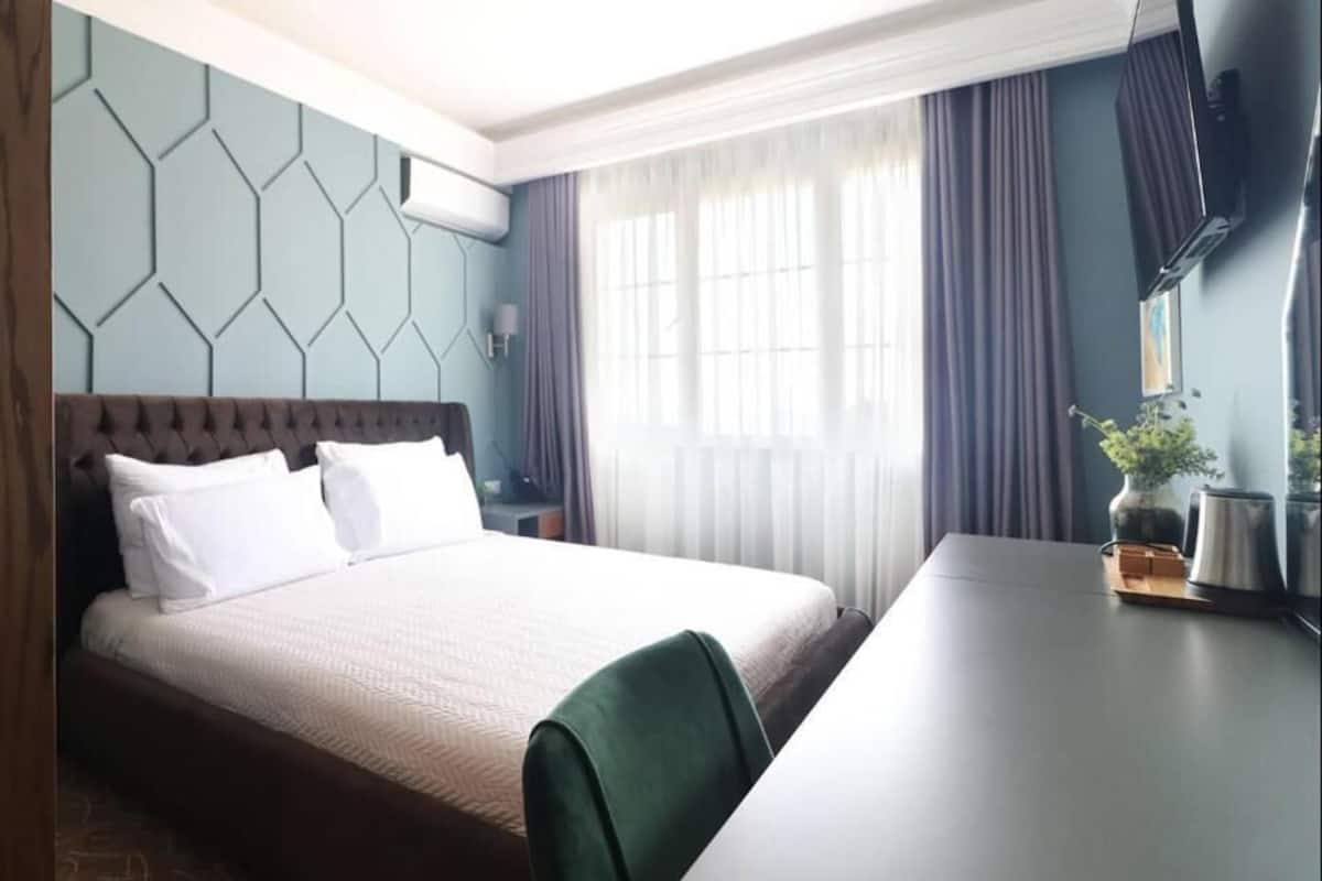 Una habitación de hotel con cama, escritorio, silla, televisor y ventana con cortinas.