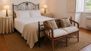 Un dormitorio ordenado con una cama, dos mesitas de noche con lámparas, un sillón de mimbre con cojines y una ventana con persianas.