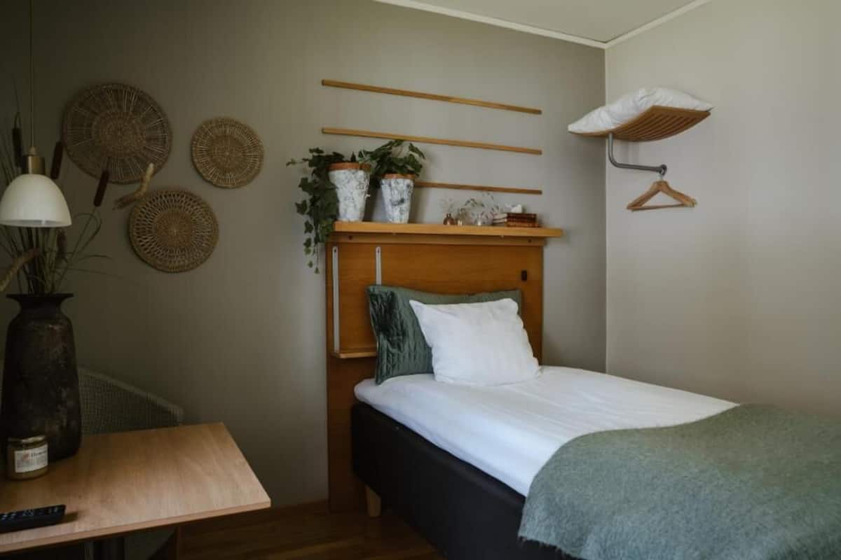 Et soveværelse med en seng, et natbord, en lampe og vægdekorationer, herunder vævede kurve og en hylde med planter.