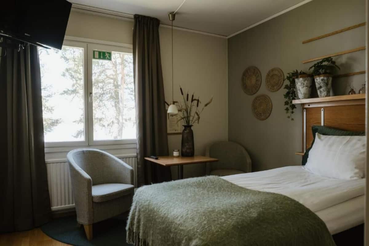 Et hotelværelse med en seng, en stol, et lille bord med en vase og en reol med planter.