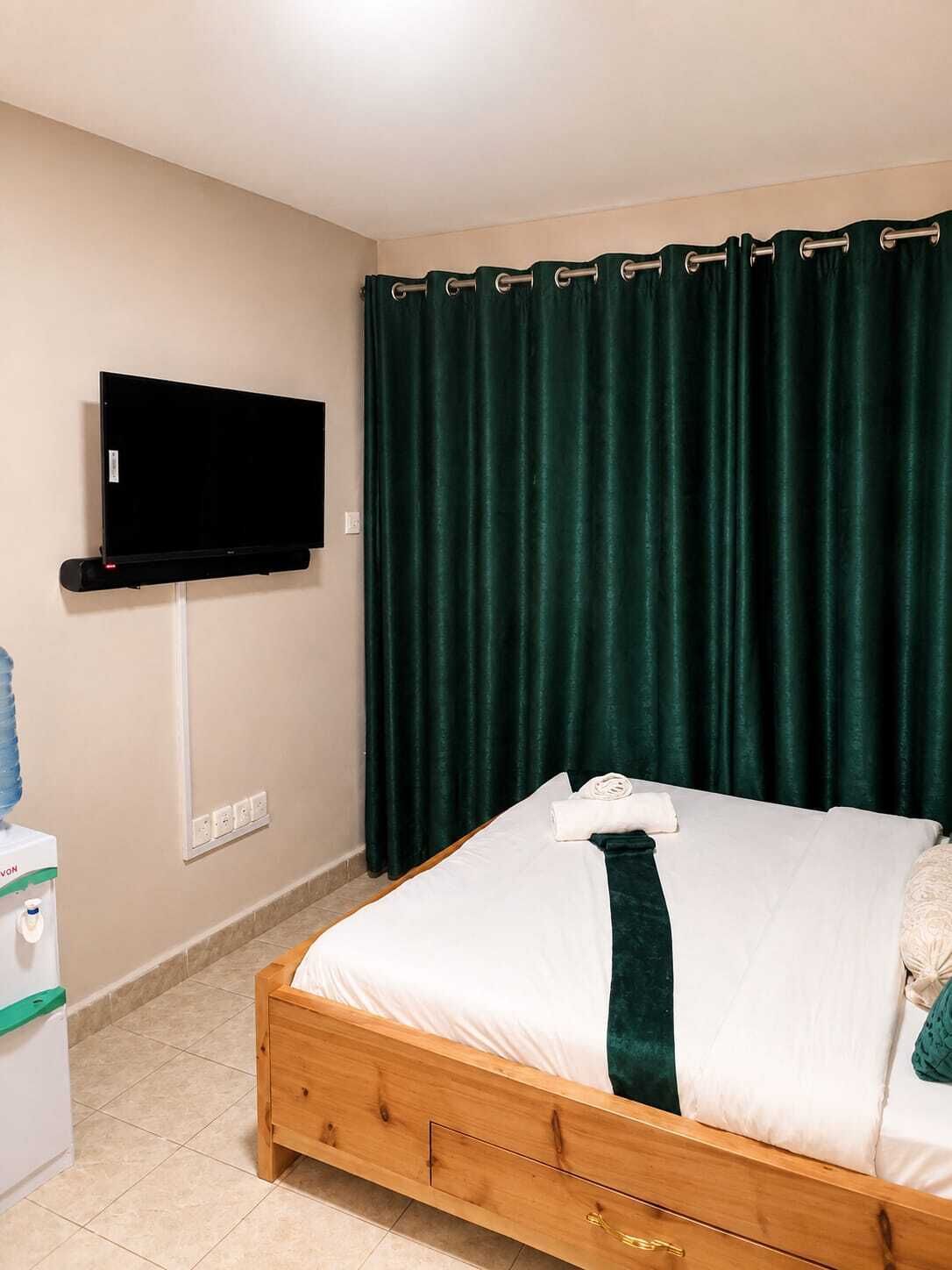 A hotel room with a bed, a flat-screen TV mounted on the wall, a water dispenser, and a green curtain.