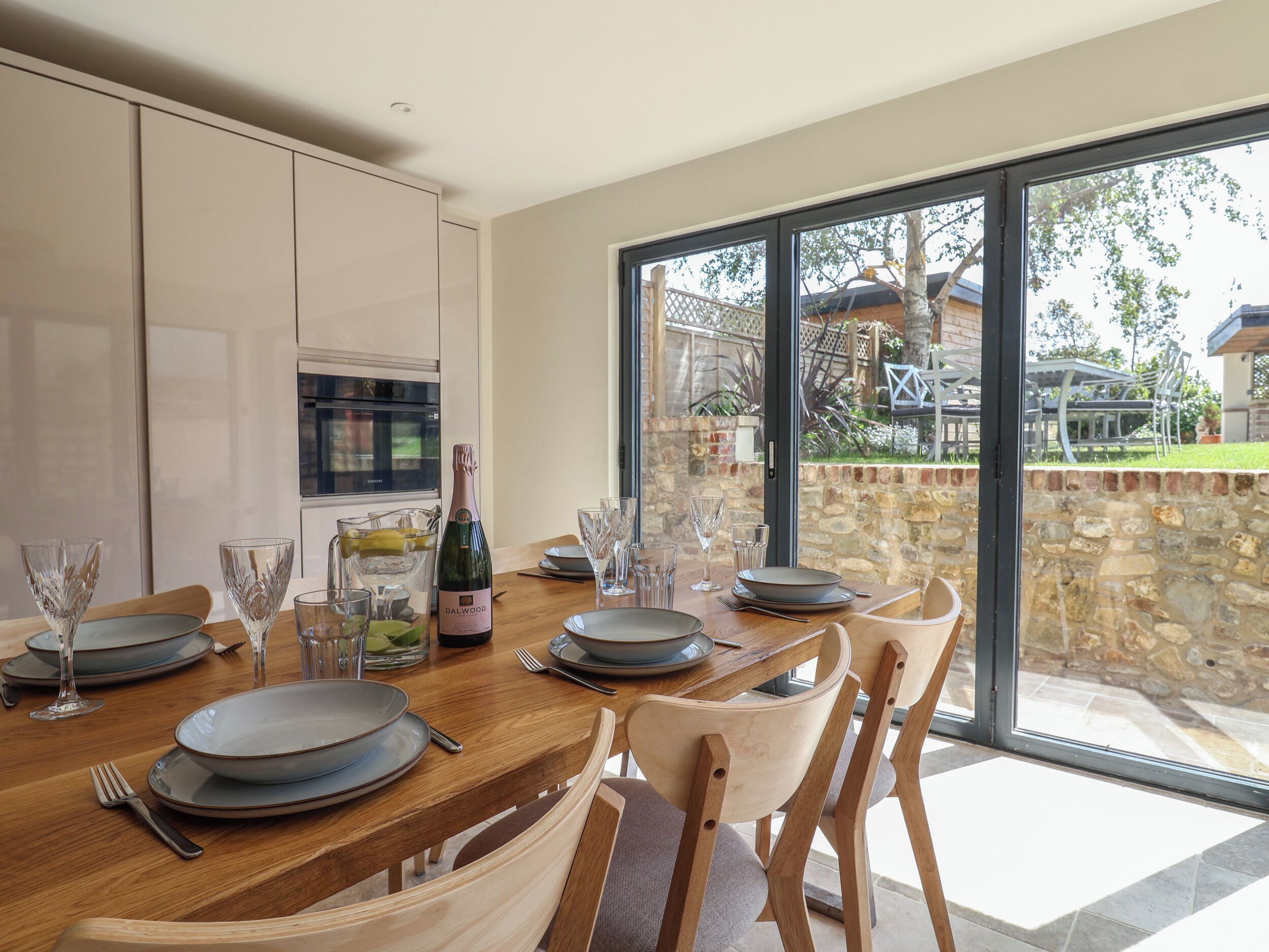 A dining area with a table set for four, a bottle of champagne, and a view of a stone wall and garden.