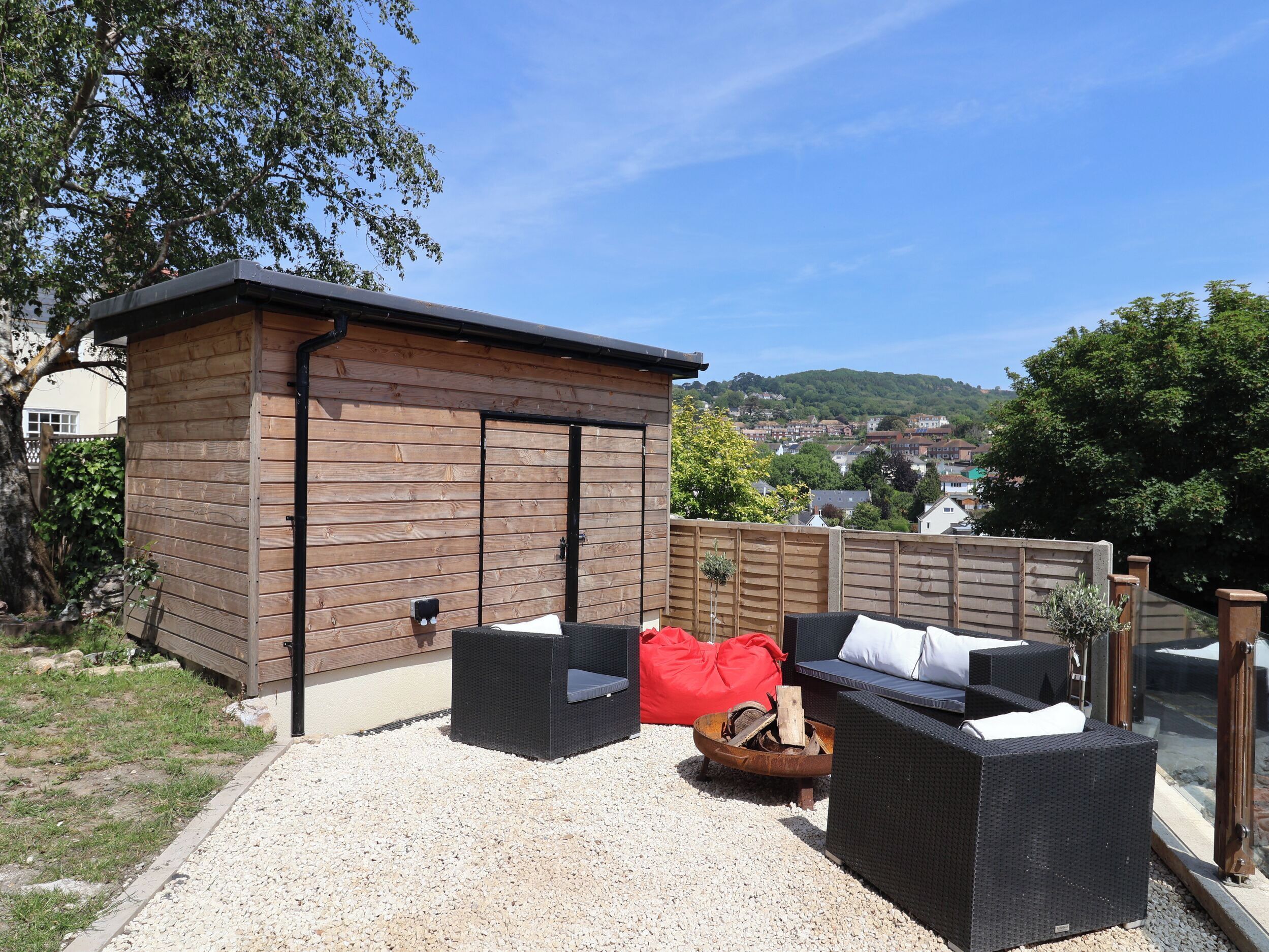 A wooden outdoor seating area with a fire pit, a red bean bag, and a view of a town in the background.