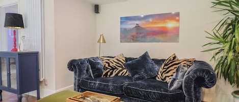 A living room with a dark velvet sofa, a green ottoman, a framed painting, a lamp, and a potted plant.