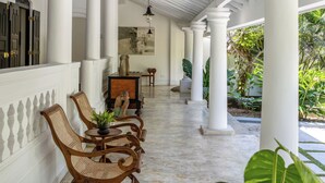 Un espacio al aire libre cubierto con columnas blancas, sillas de mimbre, una mesa con un florero y una lámpara colgante.