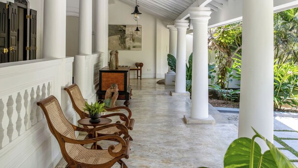 A covered outdoor area with white columns, wicker chairs, a table with a vase, and a hanging light fixture. - The Well House Galle offers a peaceful tropical villa stay in Unawatuna. (Unawatuna)