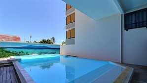 A swimming pool with a view of a tropical landscape, a wooden deck, and a modern building with balconies. - Mahana Suite (Papeete)