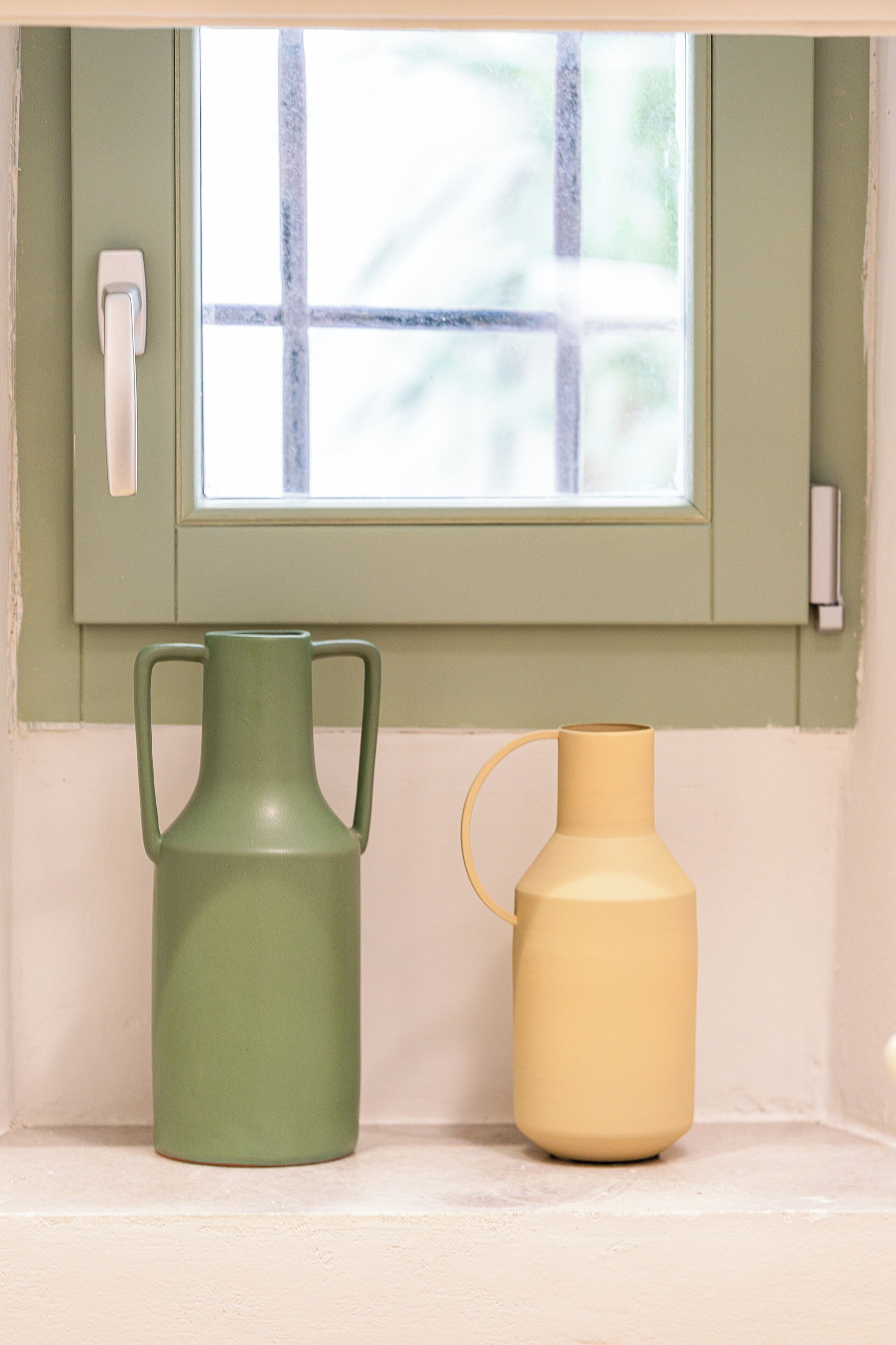 A window with a view of a balcony, a green vase, and a yellow vase on a shelf.