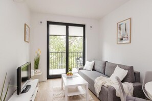 A modern living room with a grey sofa, a white coffee table, a flat-screen TV, and a balcony with a view of greenery. - ShortStayPoland Sokratesa (Warszawa)