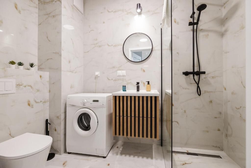 A compact bathroom with a washing machine, a toilet, a sink with a wooden vanity, a mirror, and a shower.