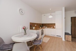 A compact kitchen with white cabinets, a wooden countertop, a round dining table with chairs, a clock on the wall, and a small rug on the floor. - ShortStayPoland Sokratesa (Warszawa)