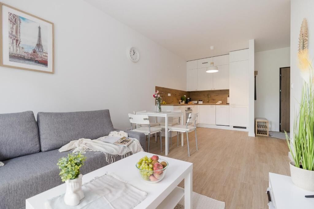 A modern living room with a grey sofa, a dining table with chairs, a kitchen area, and a clock on the wall.