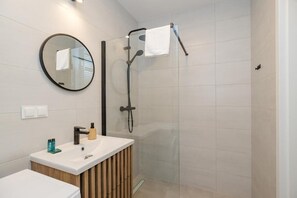 A modern bathroom with a glass shower enclosure, a round mirror, a white sink, and a wooden vanity. - ShortStayPoland Sokratesa (Warszawa)