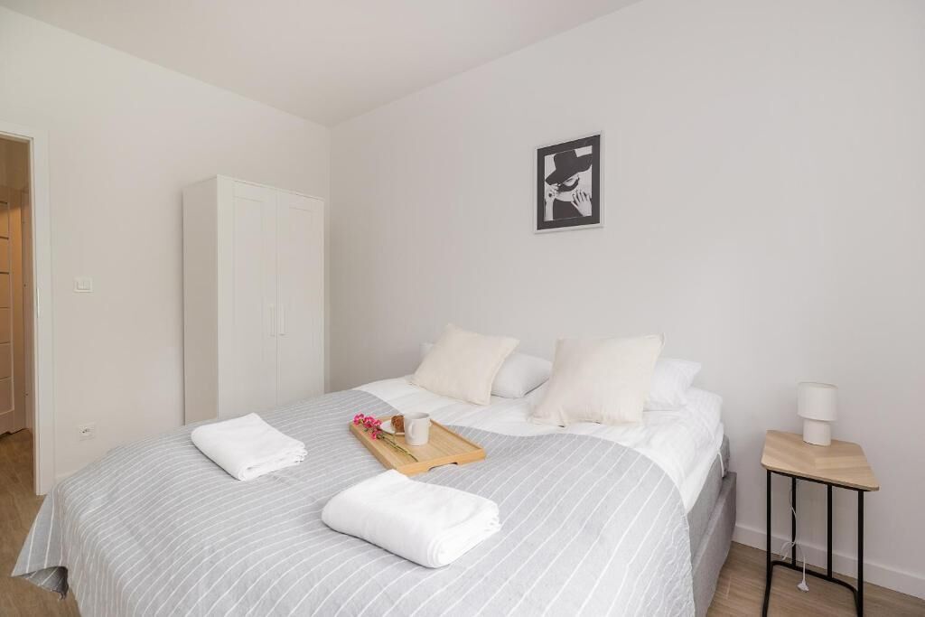 A neatly made bed with white linens, a tray with flowers and a mug, a white wardrobe, and a small bedside table with a lamp.