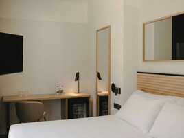 Habitación de hotel con cama, escritorio, silla, espejo, televisión y minibar.