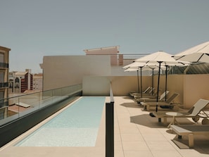A rooftop pool area with lounge chairs and umbrellas, a clear sky, and a view of buildings in the background.