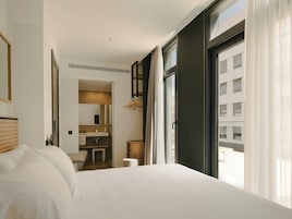 Habitación de hotel con una cama grande, un baño con espejo y lavamanos, y vista a edificios por la ventana.