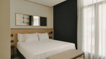 A hotel room with a bed, a bench, a large mirror, and sheer curtains.