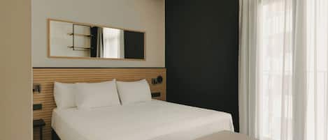 A hotel room with a bed, a bench, a large mirror, and sheer curtains.