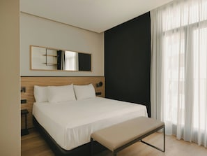 A hotel room with a bed, a bench, a large mirror, and sheer curtains.