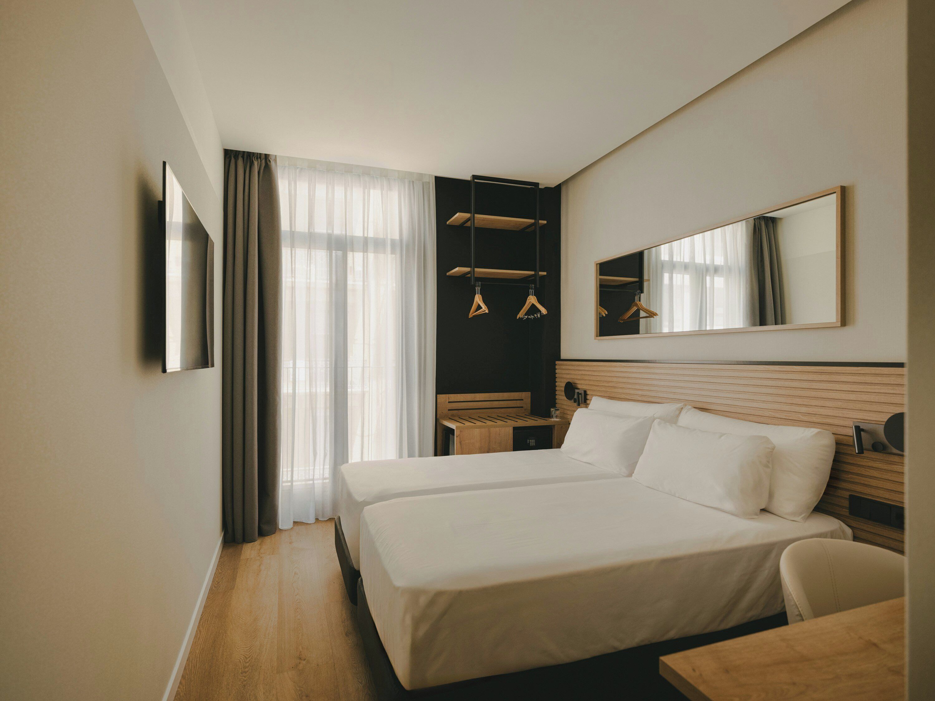 A hotel room with a double bed, a bedside table, a chair, a desk, a mirror, and a wardrobe with hanging clothes.
