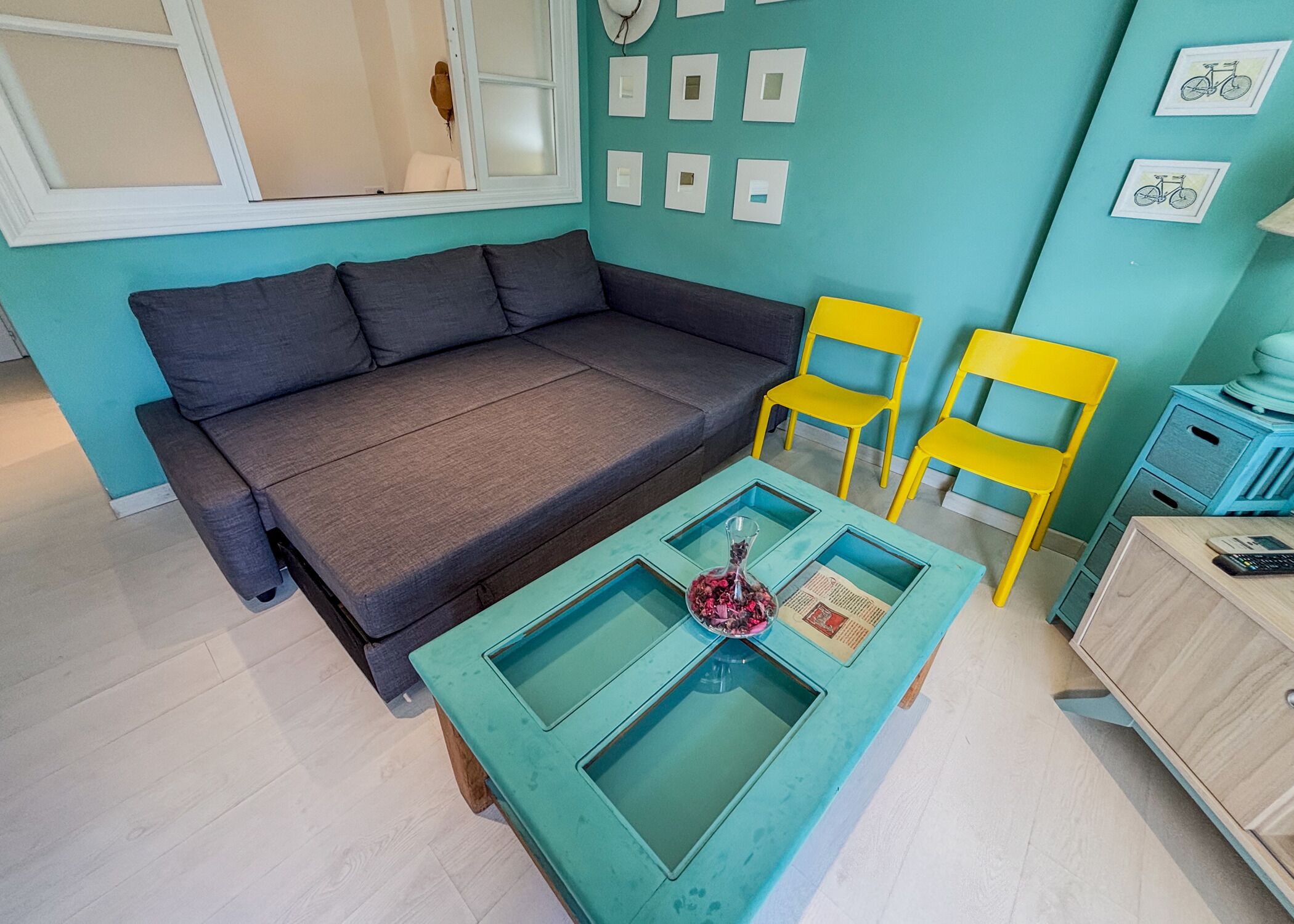 A teal-colored room with a grey sofa, two yellow chairs, a glass-top coffee table with a vase, and a small wooden cabinet.